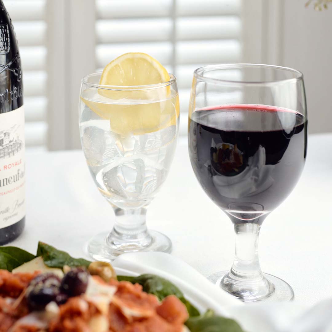 a 10.5 ounce stemmed wine glass half full of red wine next to another smaller glass of water with a lemon.